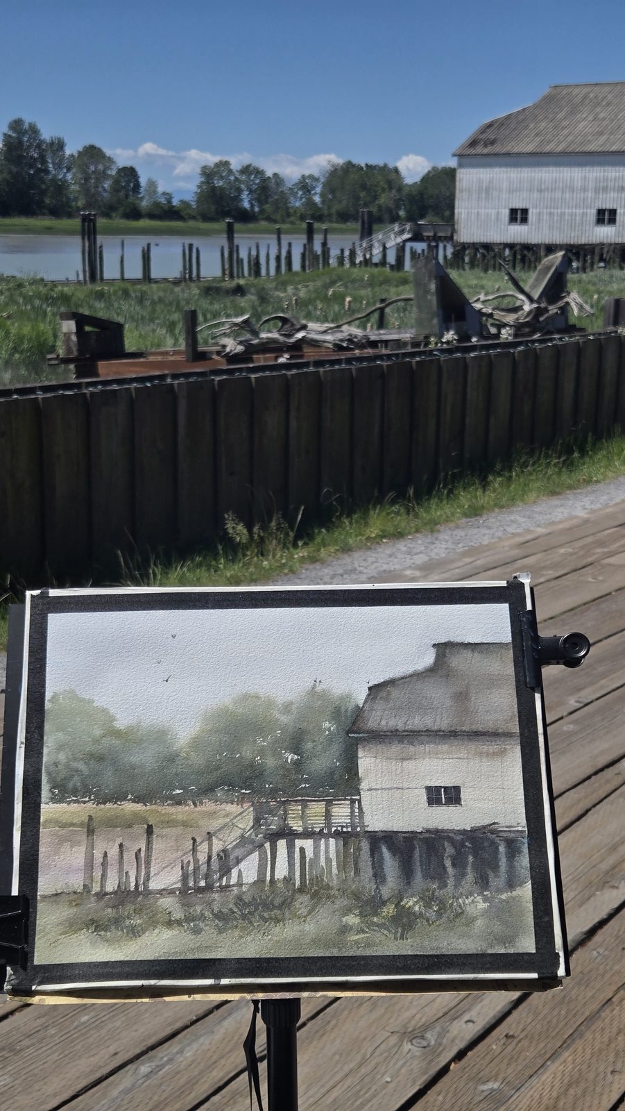 A finished framed watercolour of a heritage cannery, shown on an easel beside the real building it depicts